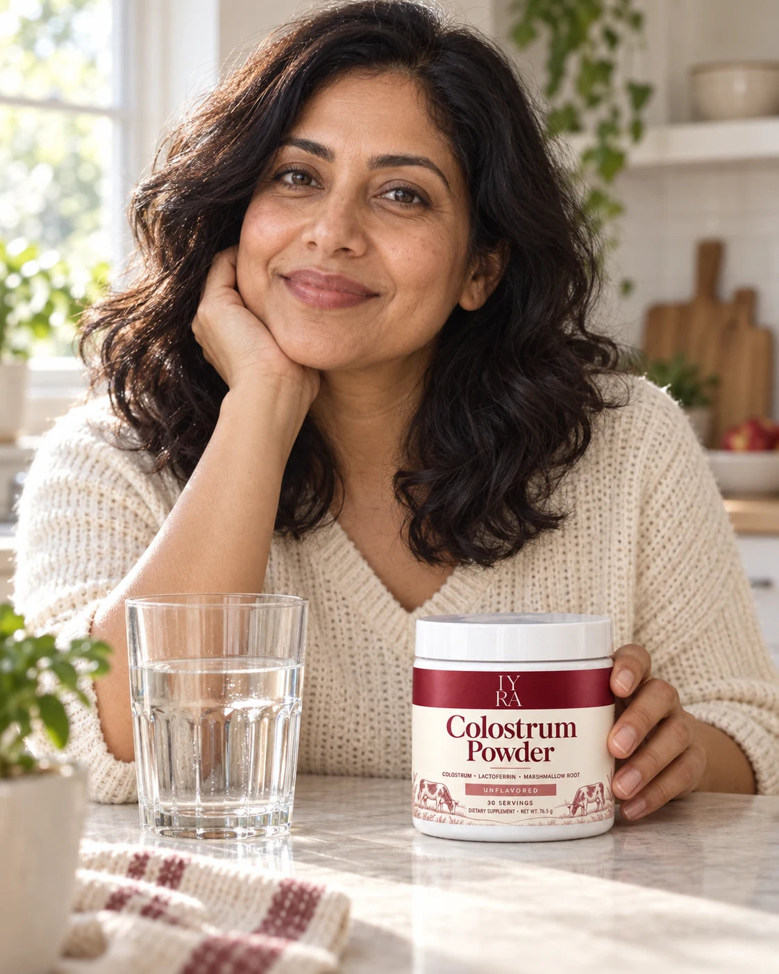 A woman sitting beside water and IYRA Colostrum Powder at a kitchen counter